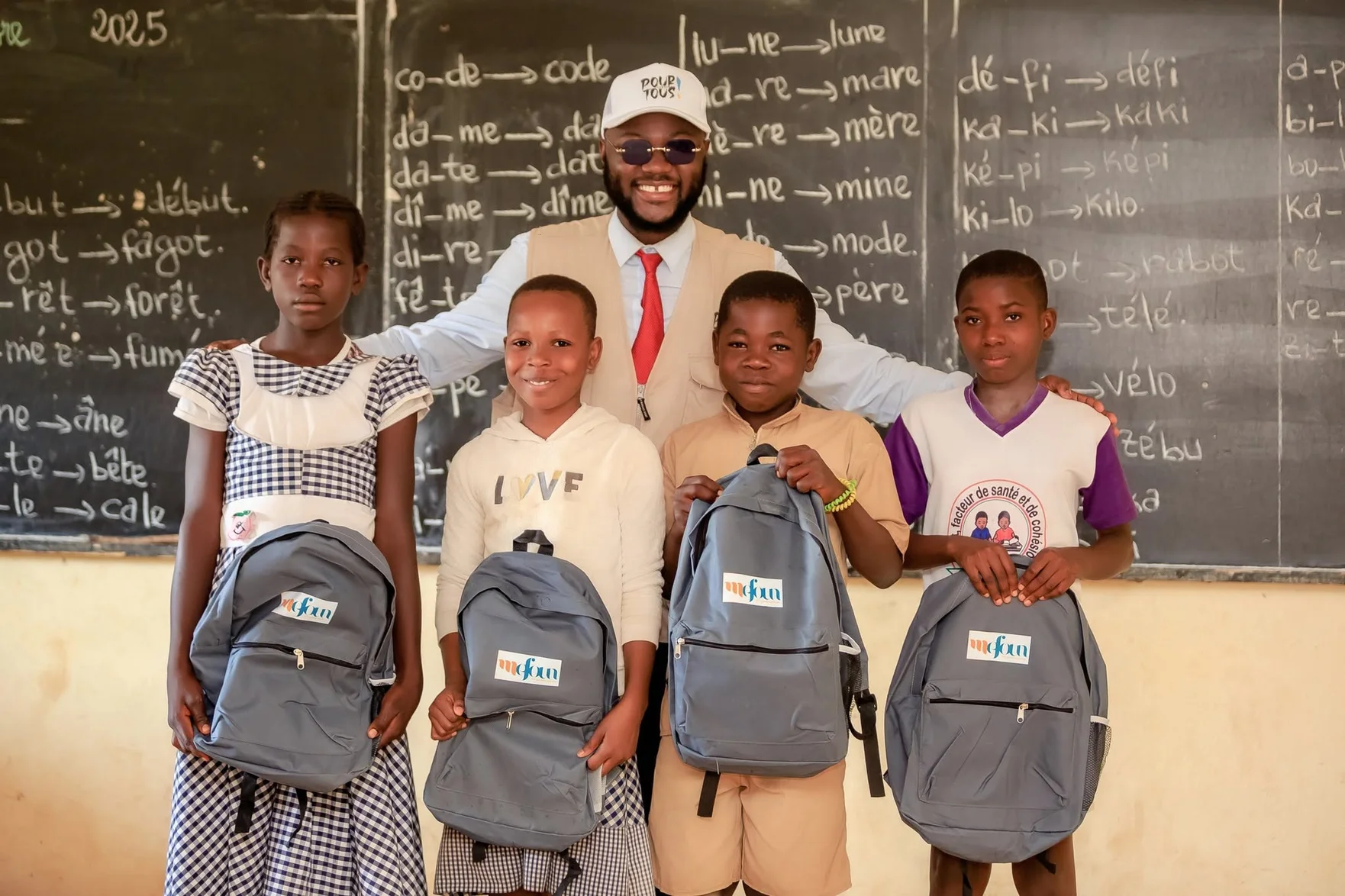 Des enfants avec leurs cartables et un membre de MEFOUN dans une salle de classe.